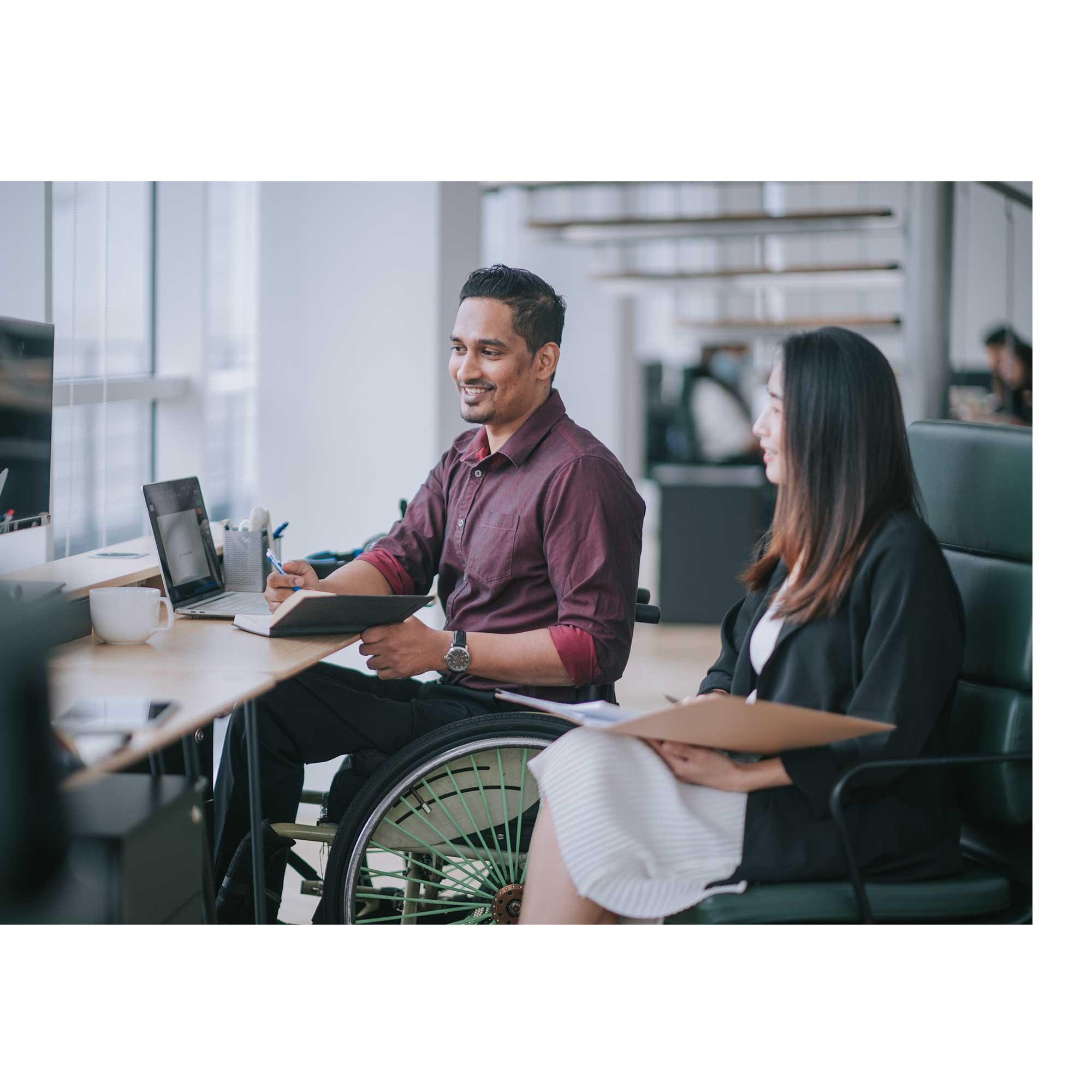 A man in a wheelchair and a woman in a desk chair sit together in an office setting, smiling and working together at a computer.