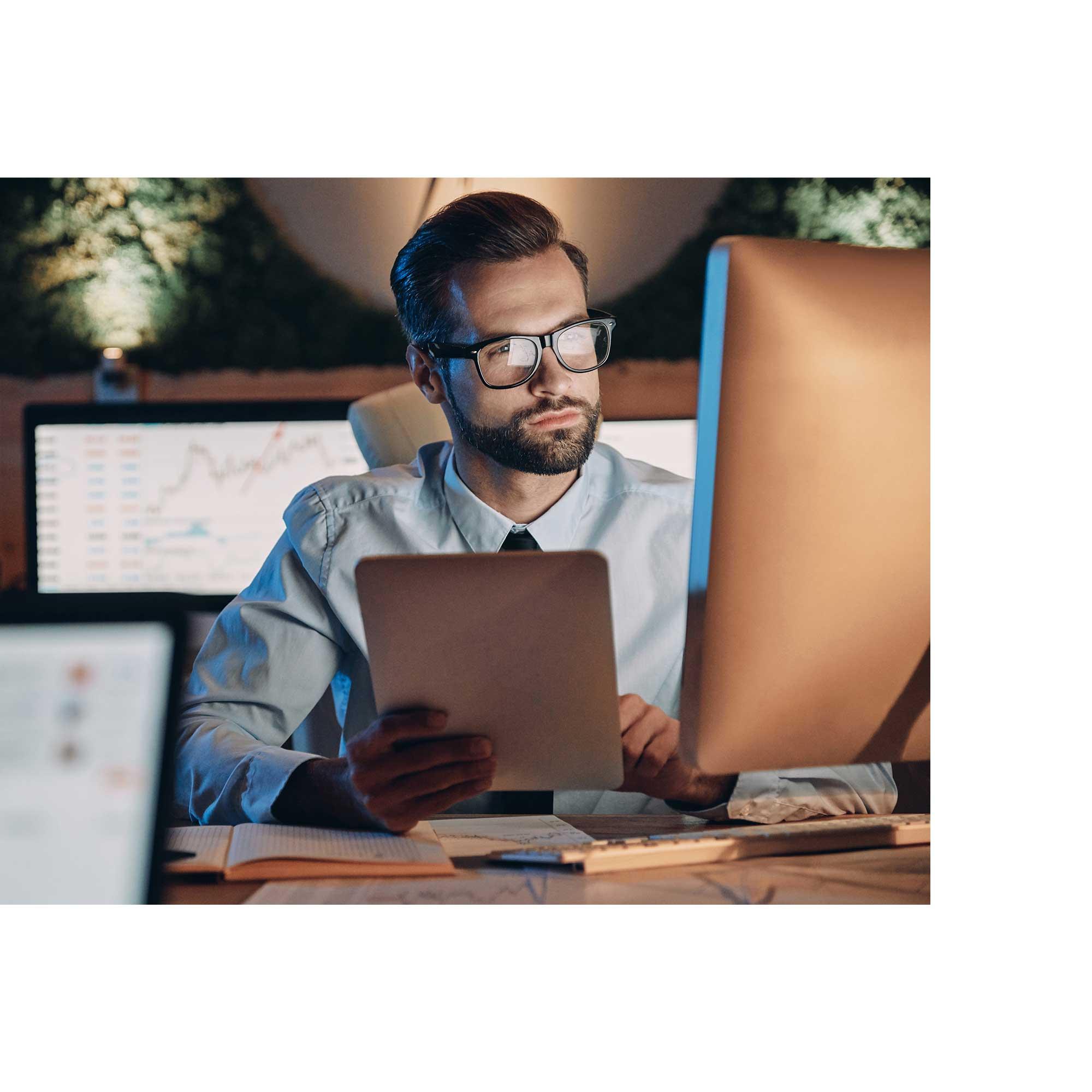 A man in glasses is sitting in front of a computer, looking at the screen. He holds a tablet in his hands. Behind him you can see data displayed on other monitors in charts.