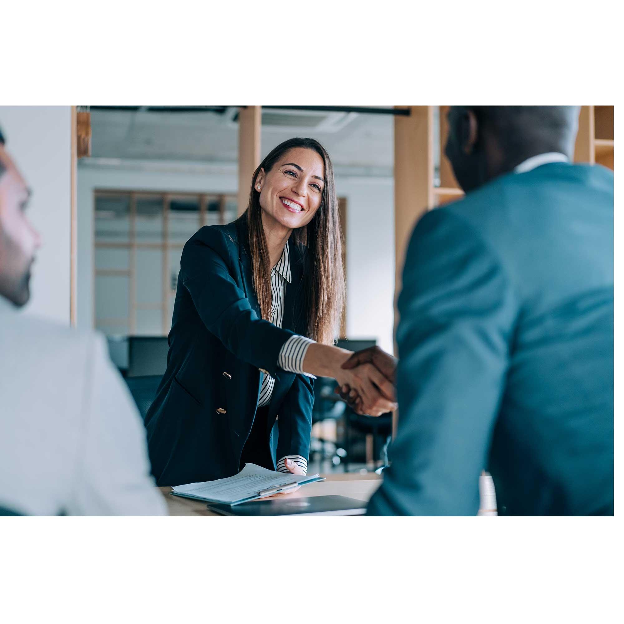 A professional in a blazer leans over a conference table to shake the hand of a man in a blazer. She is smiling in an office setting.