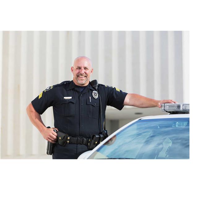 A male police officer stands with one hand on his hip, the other on the patrol car he stands next to. He is smiling at the camera.