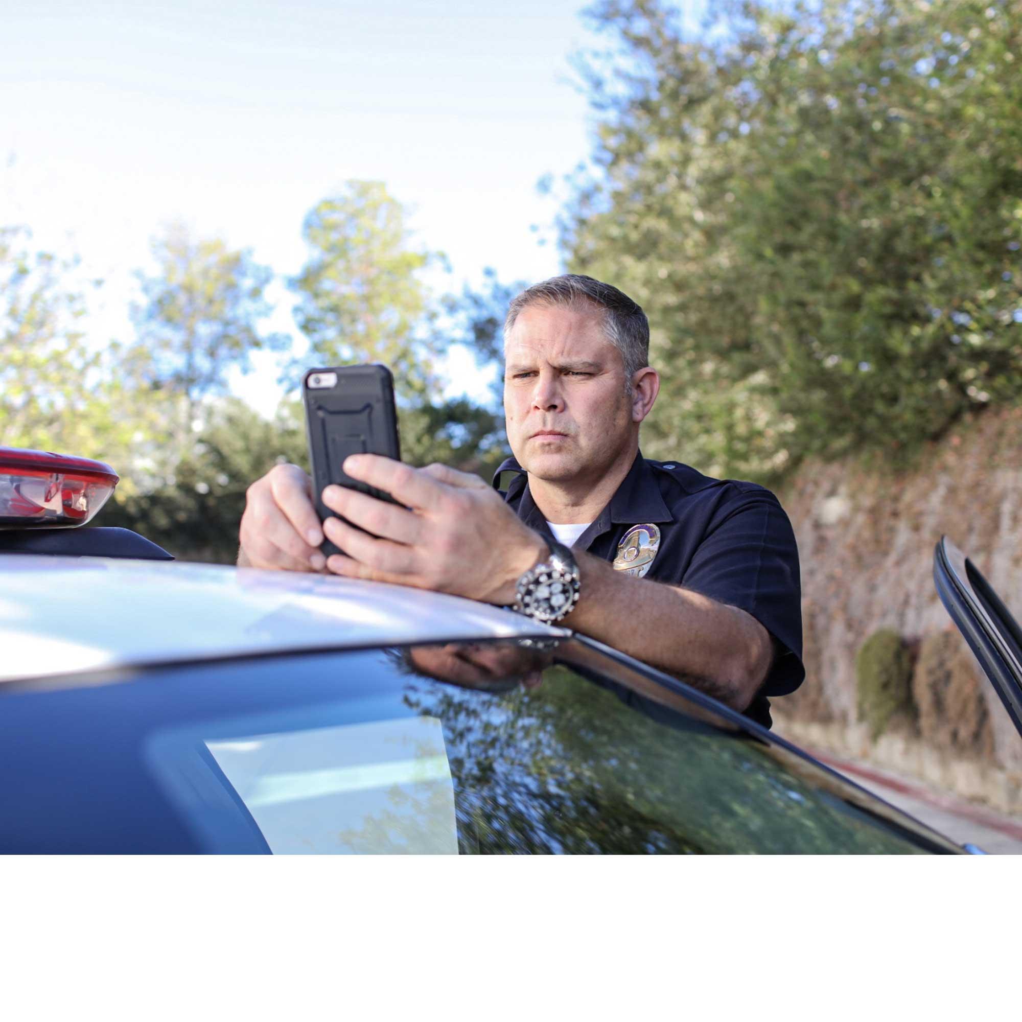 A male police officer leads on his patrol car and looks at a mobile phone with concentration.