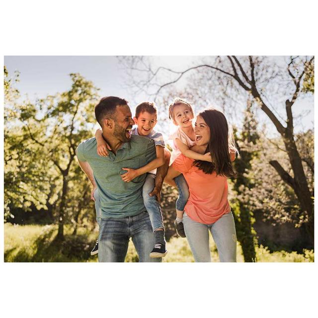 A man and woman smile and laugh as they each give a piggy back ride to small children outside with trees.