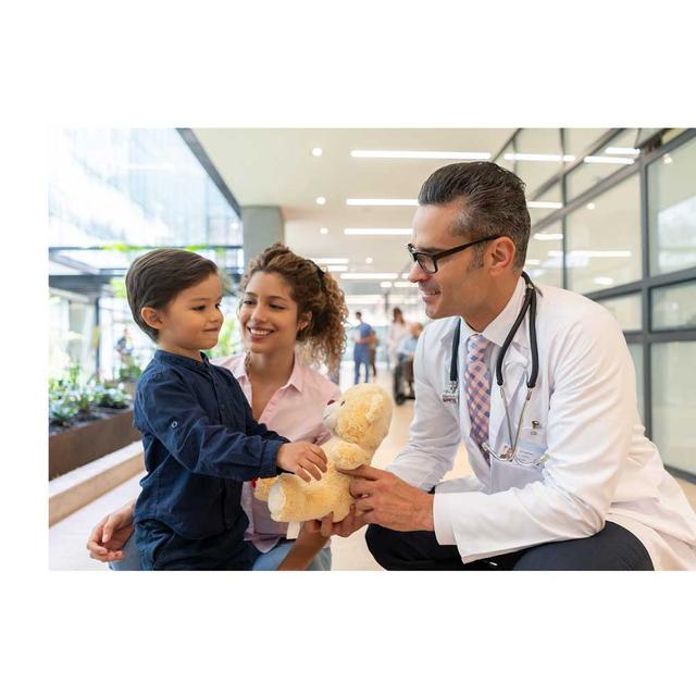 A male doctor is crouched down next to a young boy and he is handing him a yellow teddy bear. The boy's mom is next to them smiling.