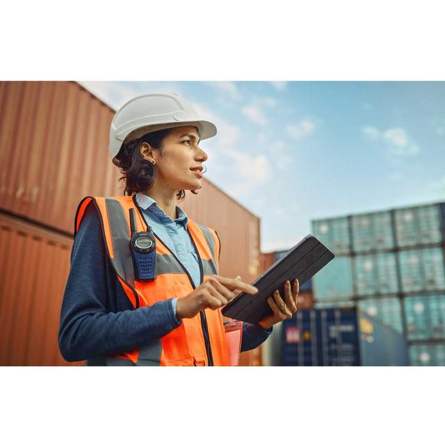 A public worker holds a tablet. She is wearing a hardhat and safety vest, standing in front of shipping containers. She looks concentrated.