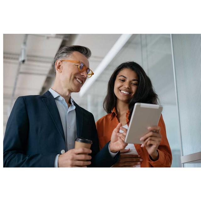 Two fashionable colleagues smile as they look together at a tablet. The man is pointing to something on the screen and holding a coffee, while the woman holds the tablet they are reading from.