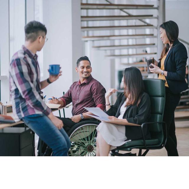 Four colleagues, one in a wheelchair, chat together at a desk in a bright and airy office setting.