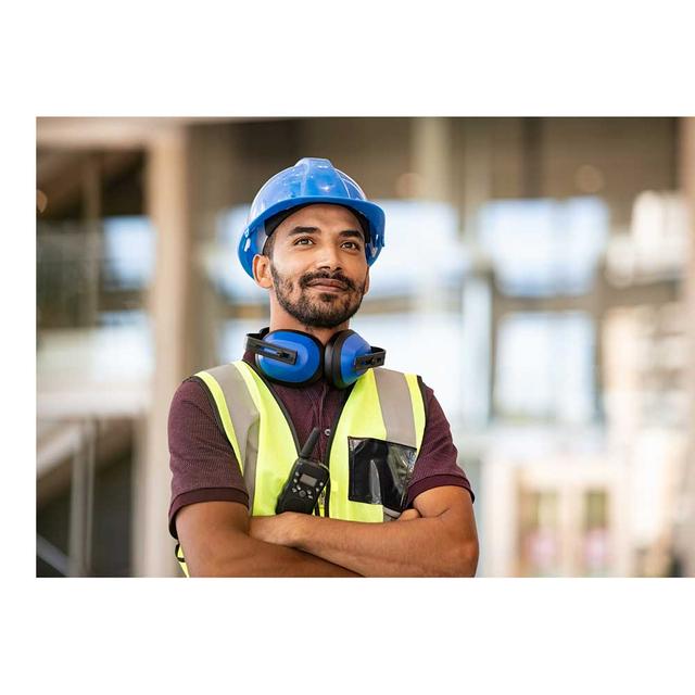 A public worker smiles at the camera. He is wearing a hardhat, safety vest and has protective ear coverings around his neck.