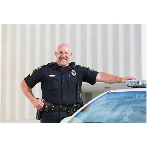 A bald male police officer stands with one hand on his hip, the other on the roof of his patrol car. He is smiling directly at the camera.