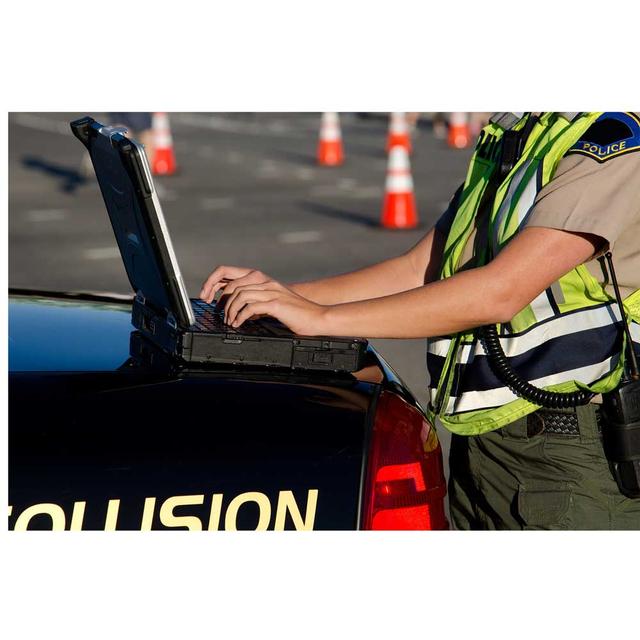 A uniformed police officer is typing on a mobile laptop, which is open on the hood of patrol car. The officer is wearing a reflective vest.