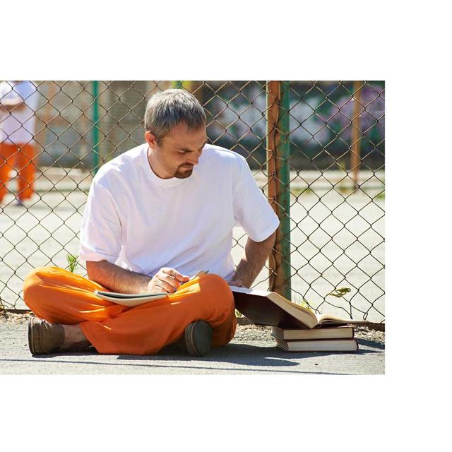 A male inmate wearing orange pants and a white shirt is sitting against a fence inside a jailyard. He has a notebook in his lap and an stack of books with an open book next to him, which he is reading.