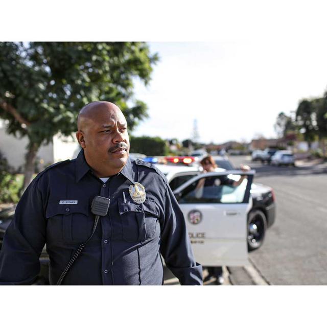 A middle aged male police officer stands in front of his patrol car, looking out of frame in concentration. Behind him, another officer is opening the patrol car door.