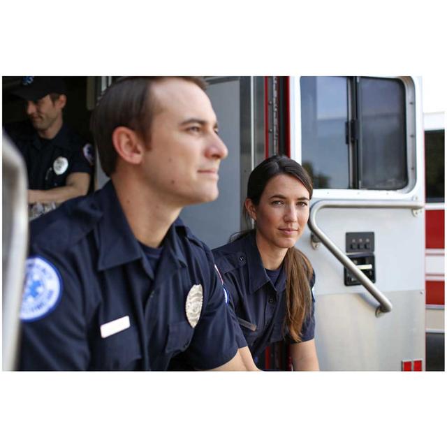 Two first responders, one man and one woman, are seated on the back of an open Ambulance. They are both smiling softly. A third first responder is visible behind them inside of the ambulance - he is also smiling.