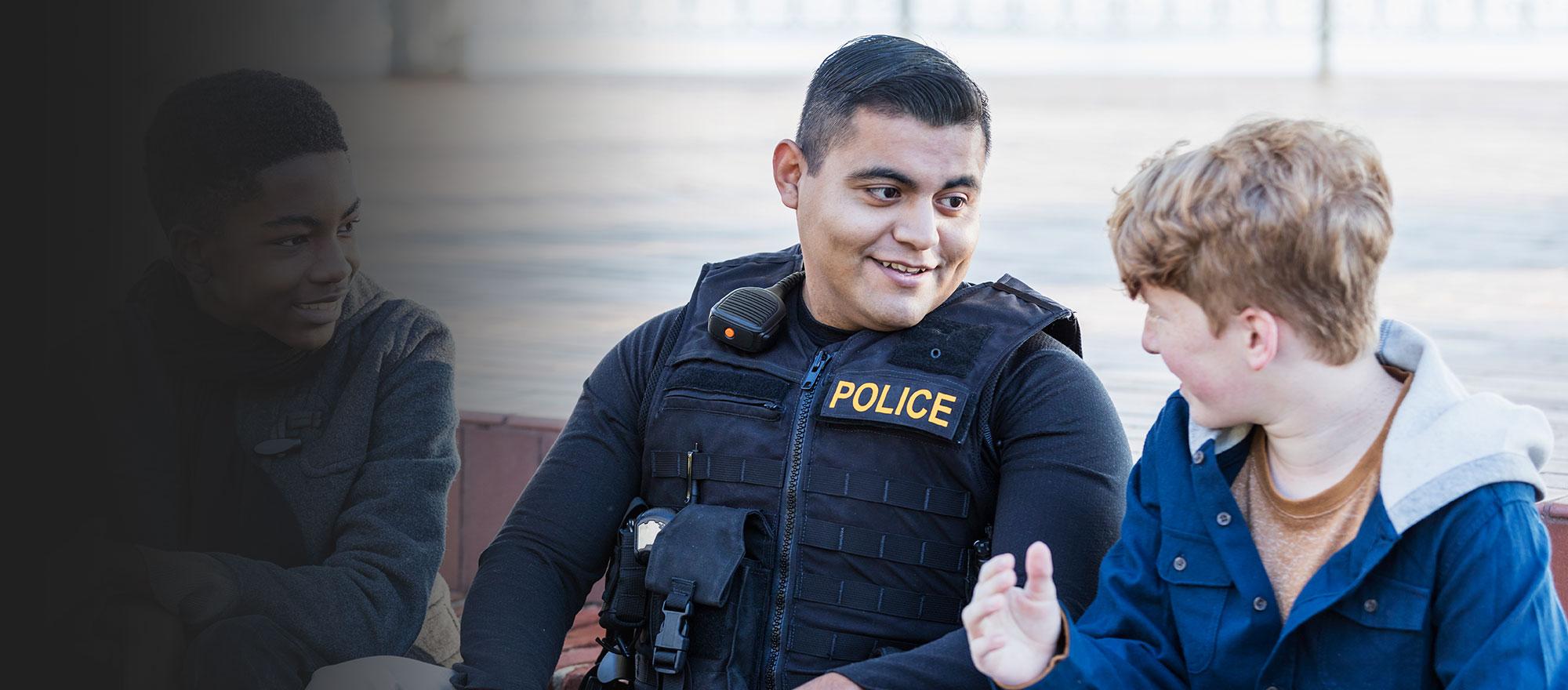 A younger appearing police officer in uniform smiles as he interacts with smiling teenage boys.