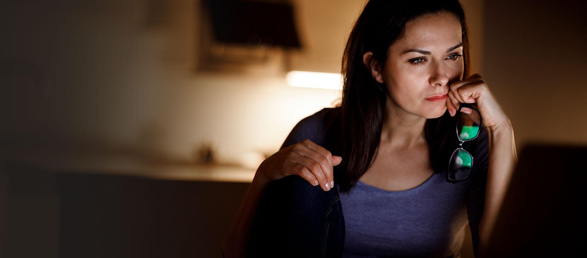 A woman sits at a desk with her eyeglasses in her hand, looking at something with focus.