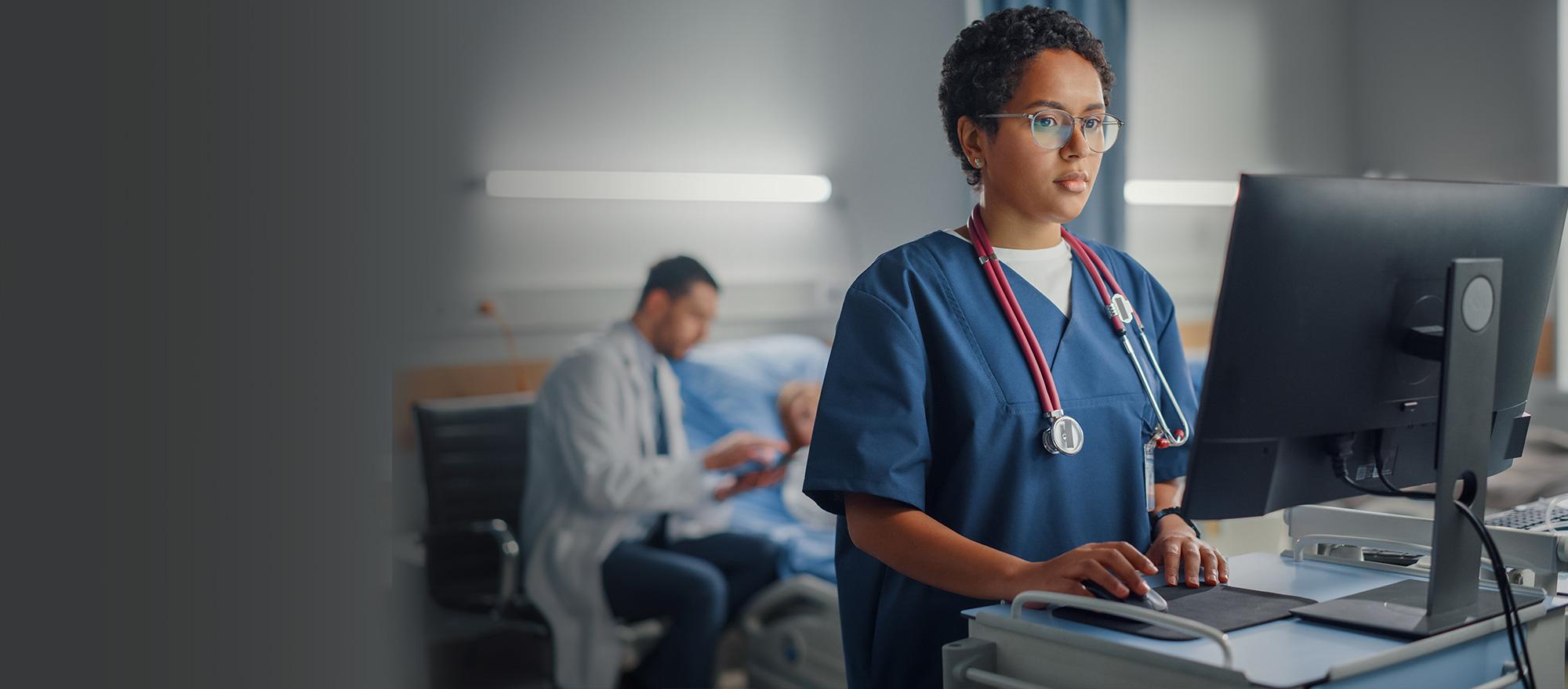 A female nurse in scrubs stands at a desktop station in a hospital room, using a computer mouse. Behind her, a doctor in a while coat consults a medical chart with a patient in the hospital bed.