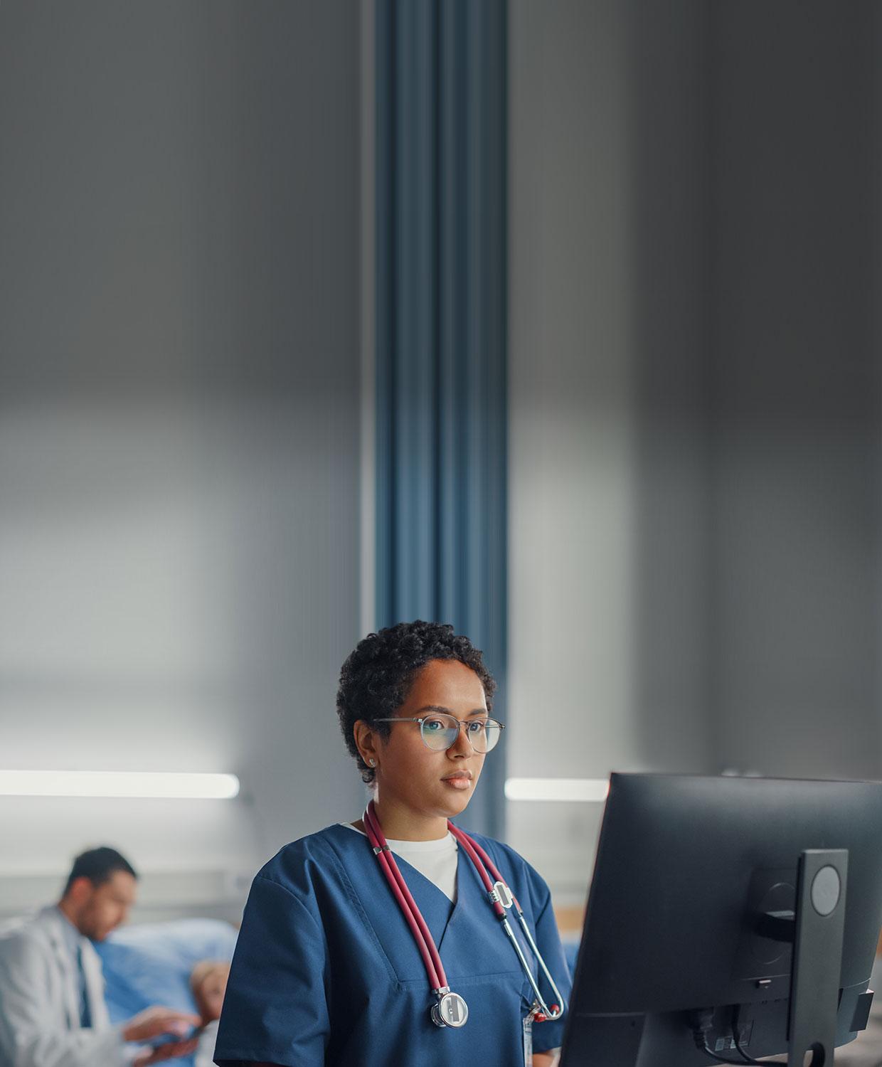 A female nurse in scrubs stands at a desktop station in a hospital room, using a computer mouse. Behind her, a doctor in a while coat consults a medical chart with a patient in the hospital bed.