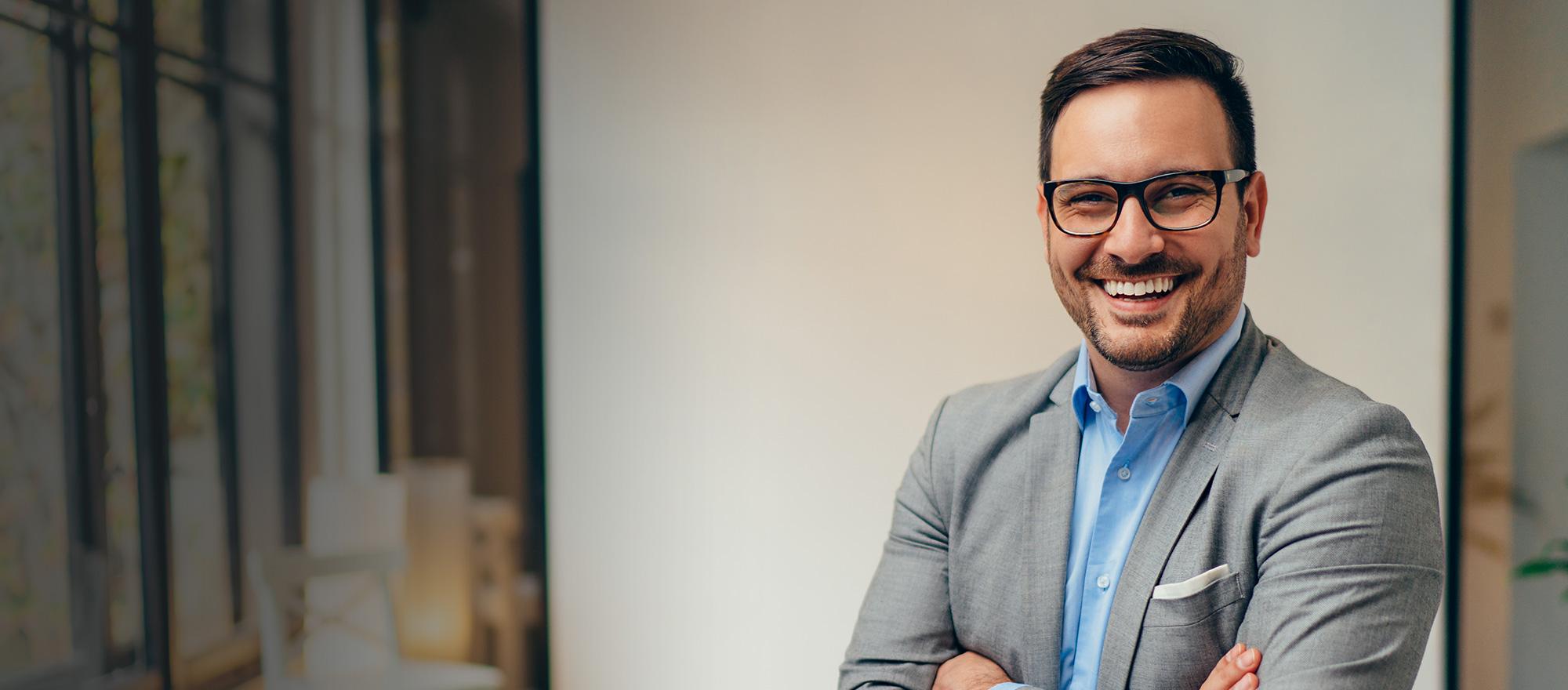 A middle aged man in glasses and business suit smiles at the camera in an office setting.