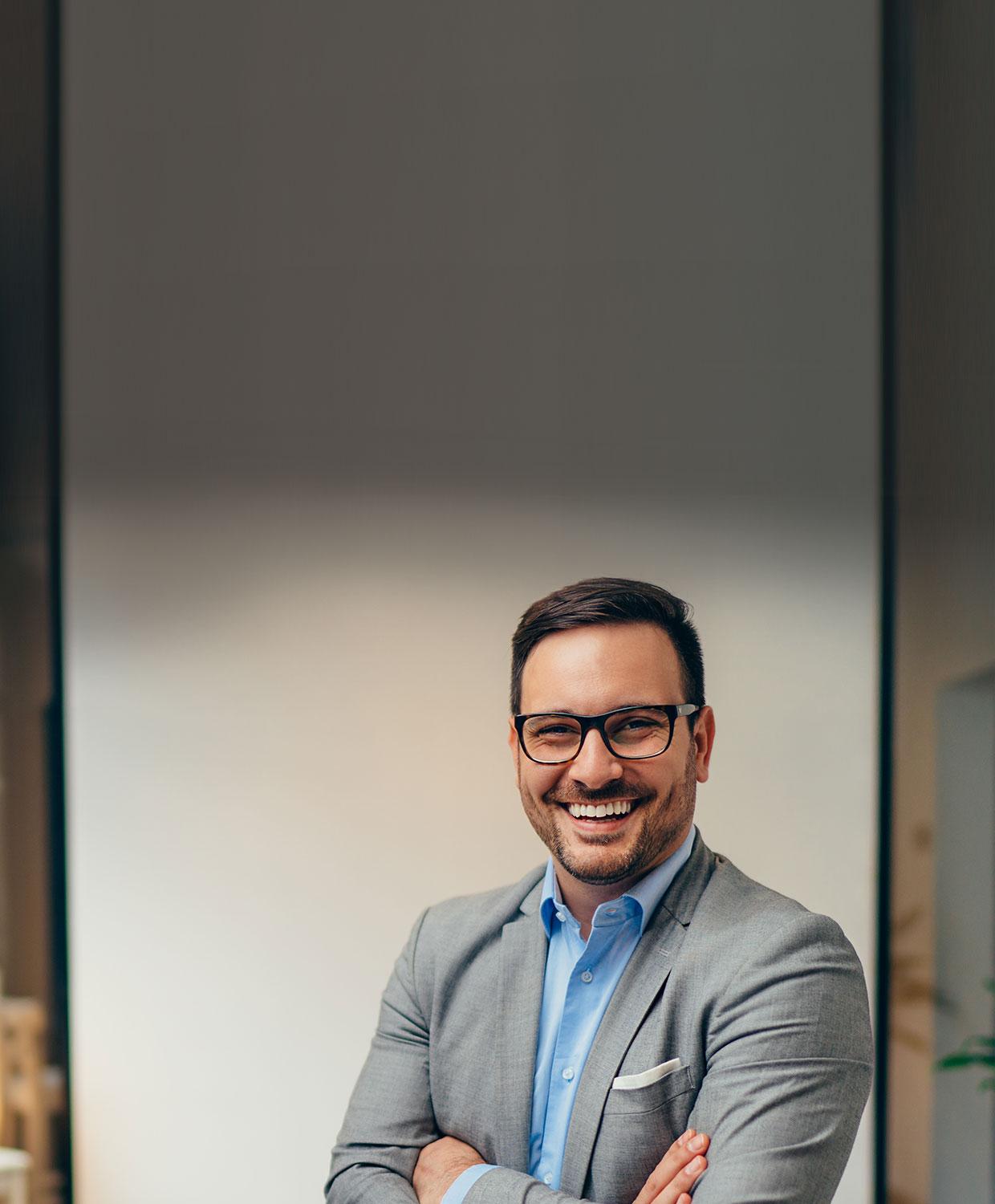 A middle aged man in glasses and business suit smiles at the camera in an office setting.