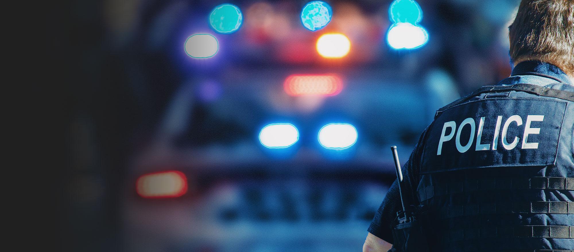 A mobile optimized version of an image - in the foreground, a uniformed police officer faces away from the camera so the back of his vest is visible, which reads "Police". In the background we see a police car with it's patrol lights on, blurred slightly.