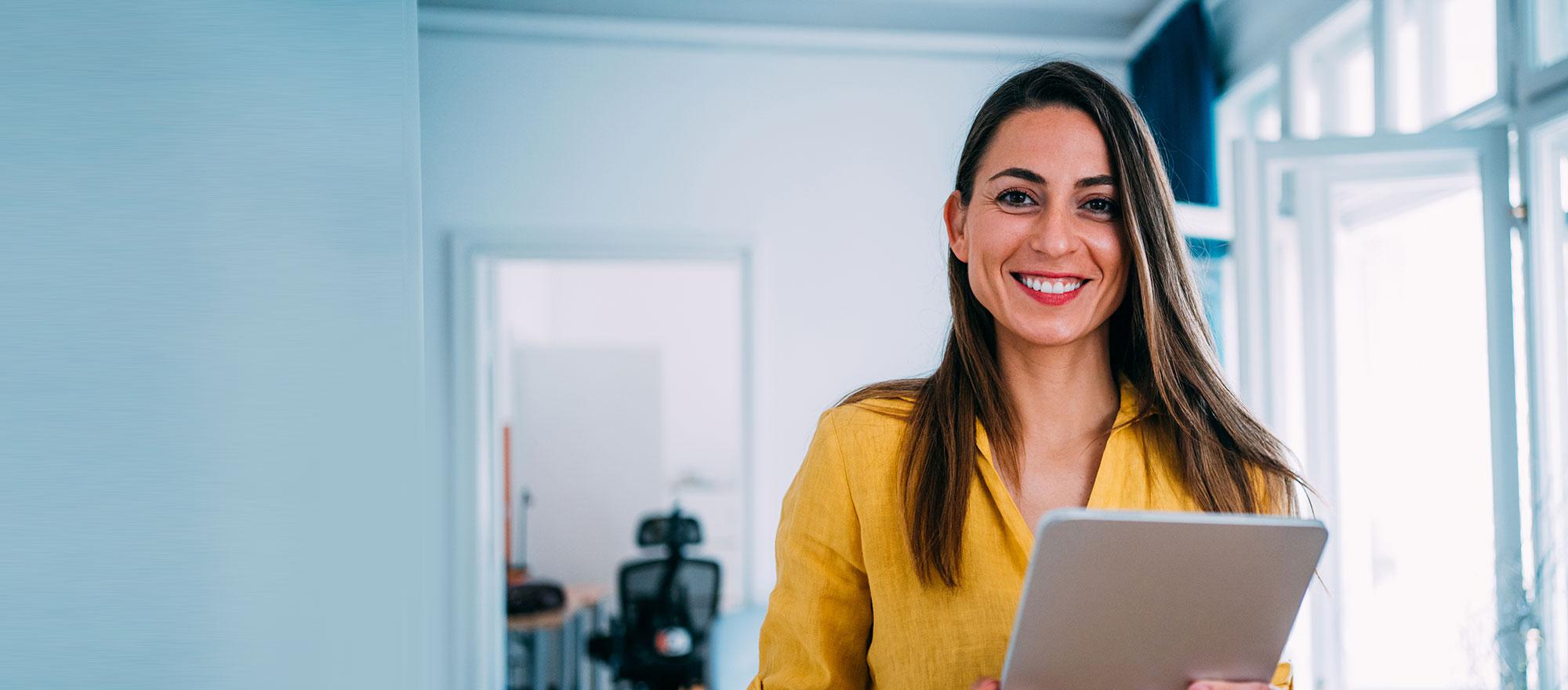 A woman in holds a stack of papers and smiles in an office setting.