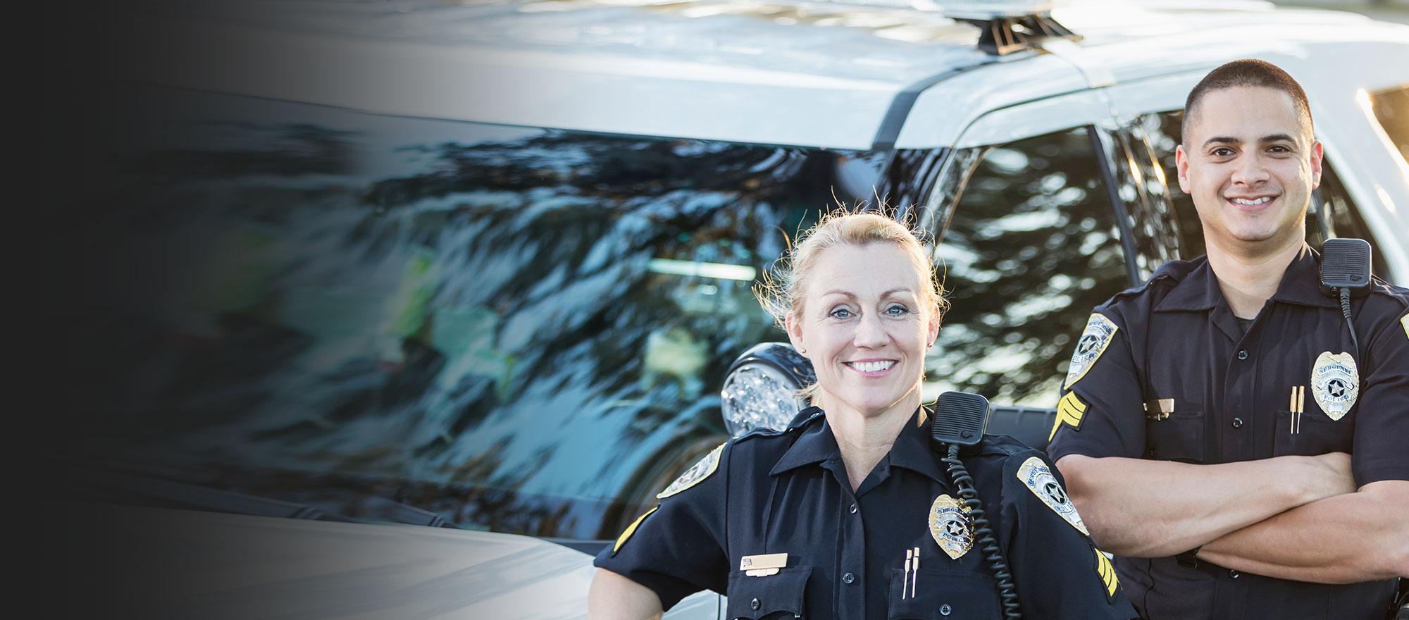 A male and female law enforcement officer stand, smiling in front of a patrol car.