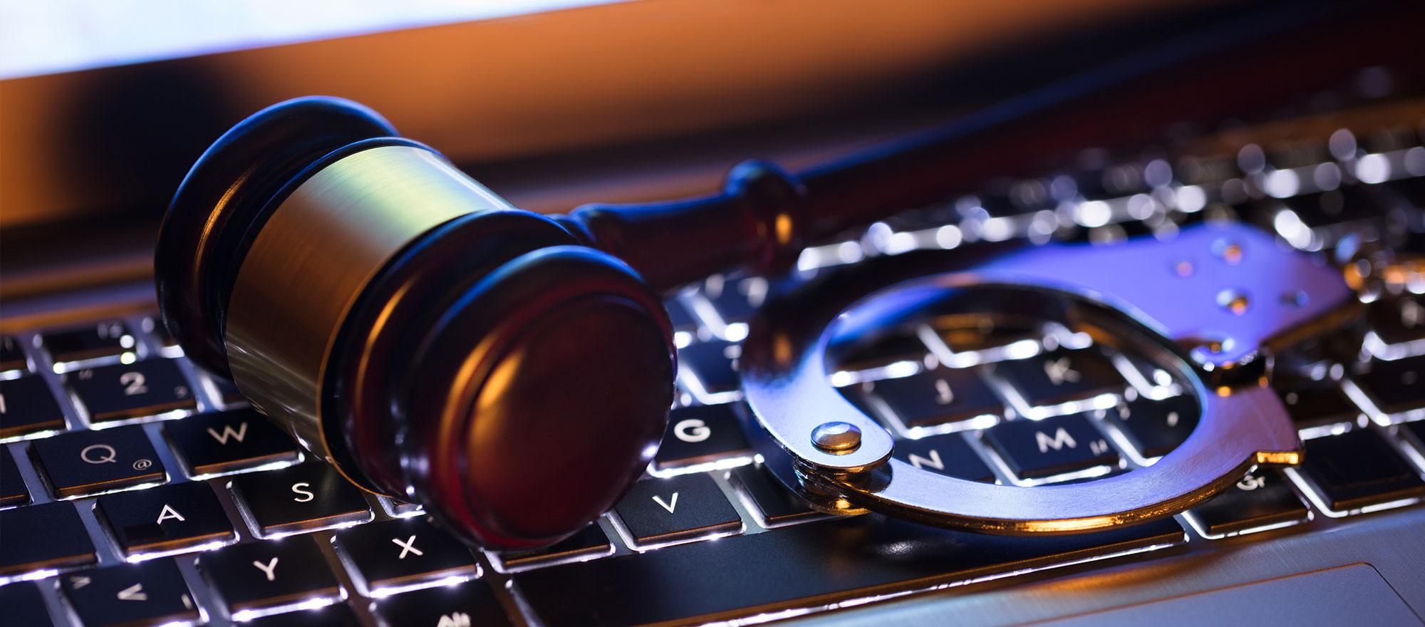 A mobile optimized image of a closeup photo of a gavel and handcuffs laid overtop the keyboard of a laptop.