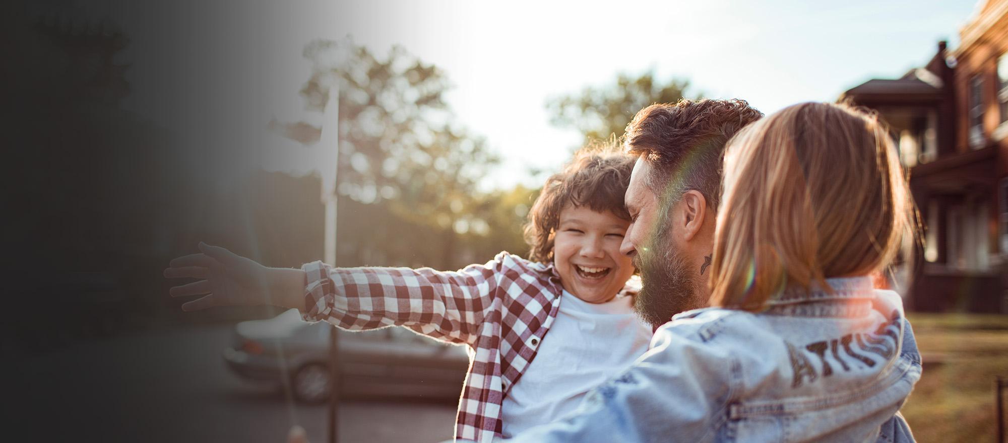 A man holds two children in his arms outdoors. They are smiling, and the children have their arms open wide.