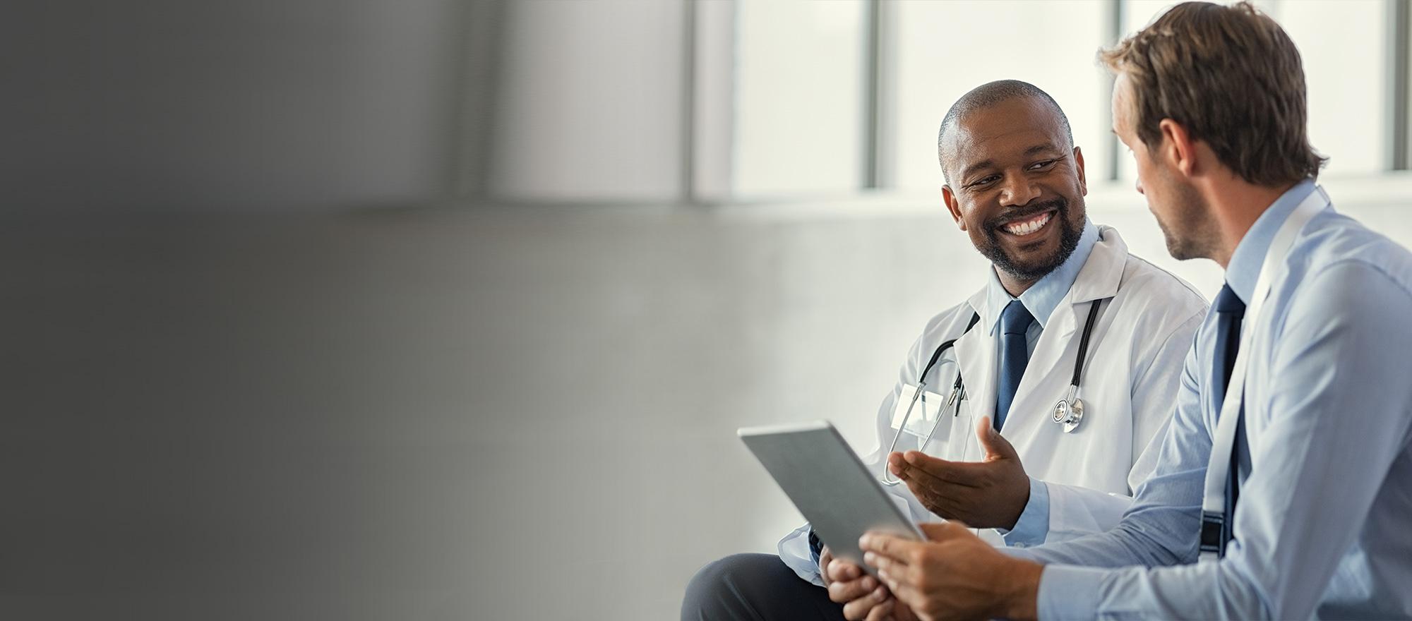 Two male doctors sit together in conversation, smiling while one holds a tablet.