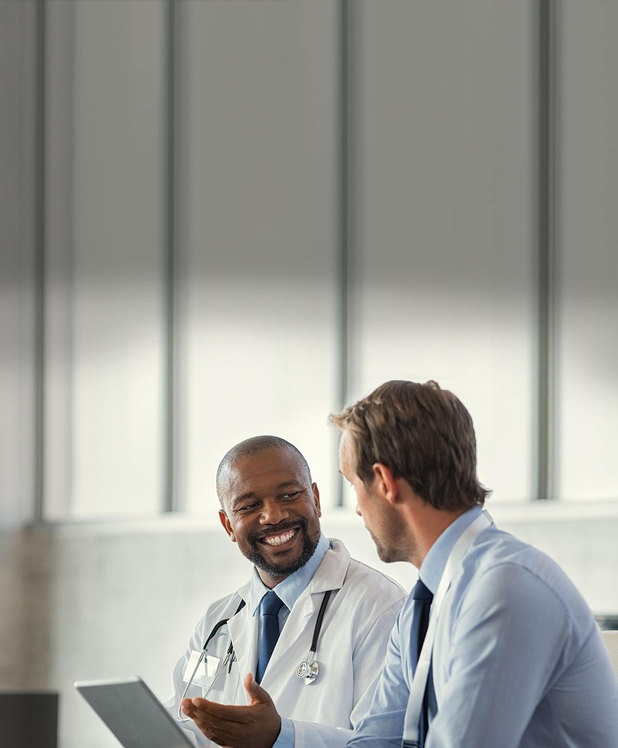 Two male doctors sit together in conversation, smiling while one holds a tablet.