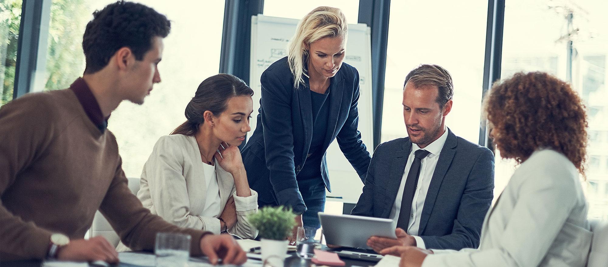 A diverse group of office workers consult a laptop screen - some are seated and others are standing, all appearing very concentrated.