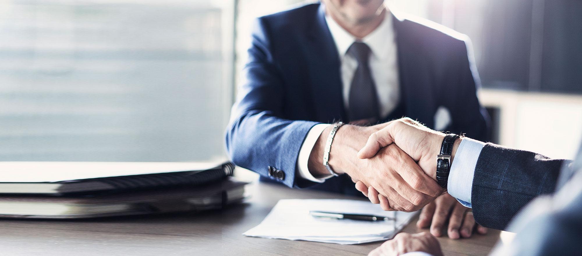 Two suited individuals shake hands across a conference room table.