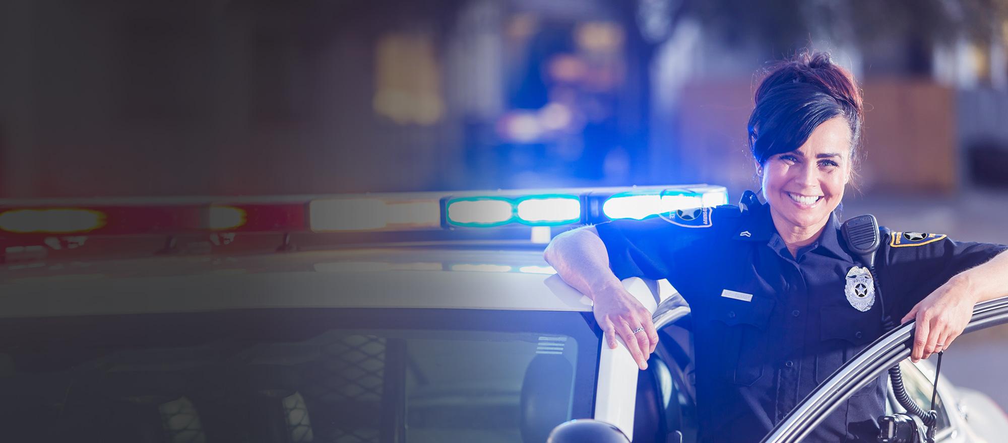 A female public safety law enforcement officer stands in the driver's door of her patrol car, smiling.