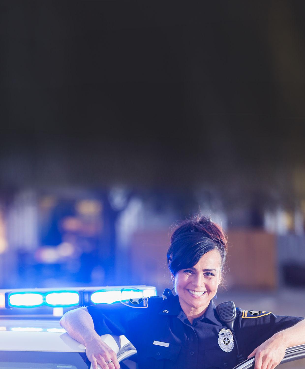 A female public safety law enforcement officer stands in the driver's door of her patrol car, smiling.