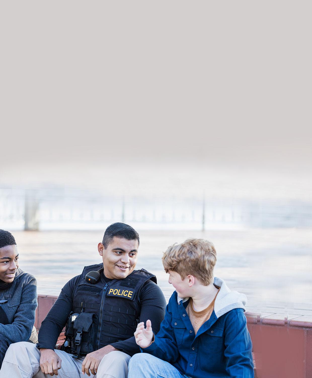 A younger appearing police officer in uniform smiles as he interacts with smiling teenage boys.