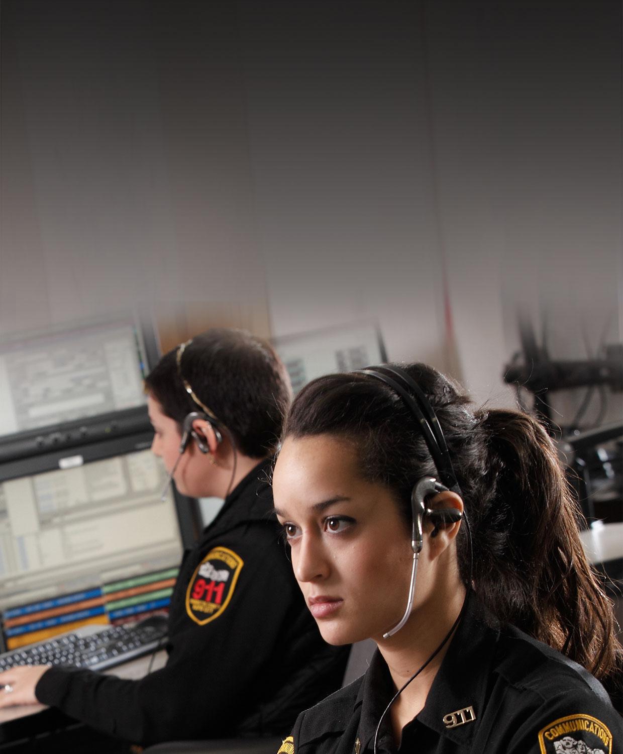 A mobile optimized version of two female telecommunicators wearing headsets are typing on computers. Multiple computer screens display CentralSquare's CAD solution.