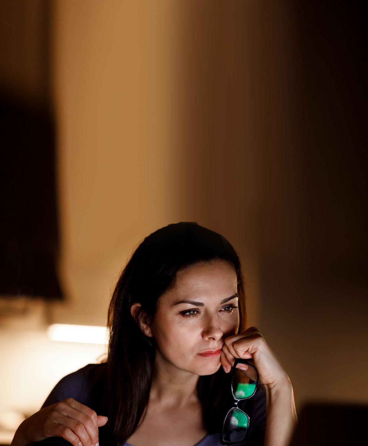 A woman sits at a desk with her eyeglasses in her hand, looking at something with focus.