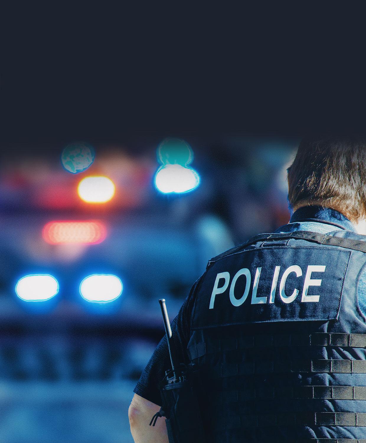 A mobile optimized version of an image - in the foreground, a uniformed police officer faces away from the camera so the back of his vest is visible, which reads "Police". In the background we see a police car with it's patrol lights on, blurred slightly.