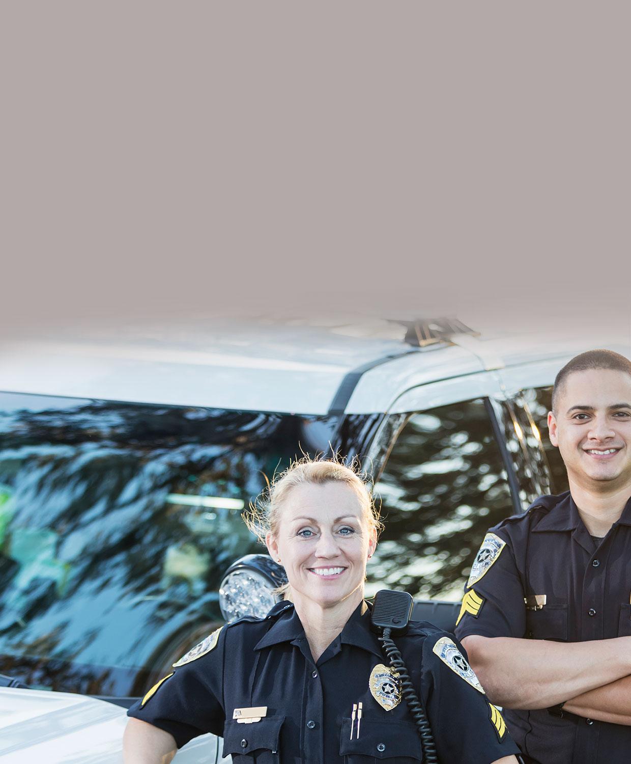 A male and female law enforcement officer stand, smiling in front of a patrol car.