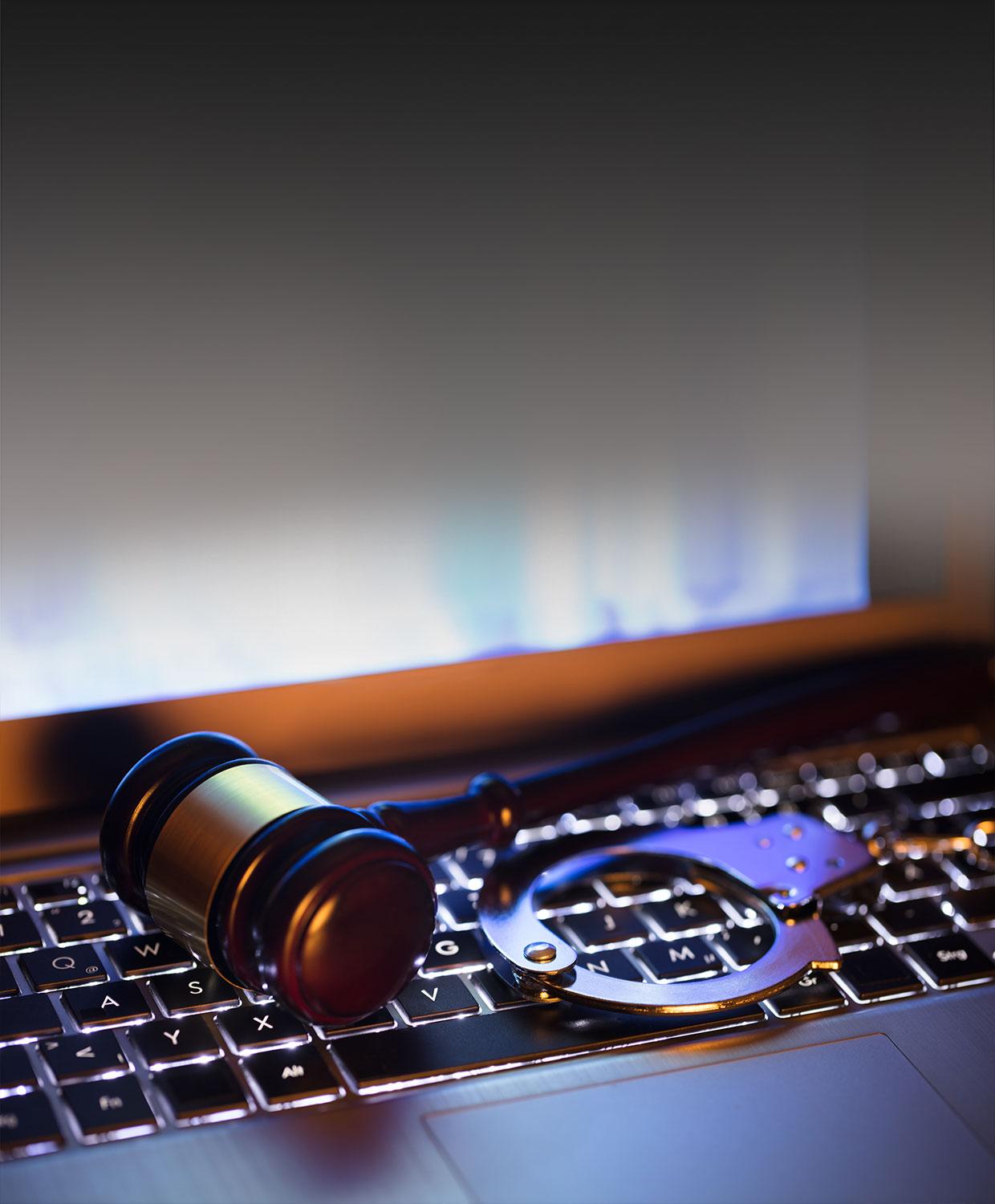 A mobile optimized image of a closeup photo of a gavel and handcuffs laid overtop the keyboard of a laptop.