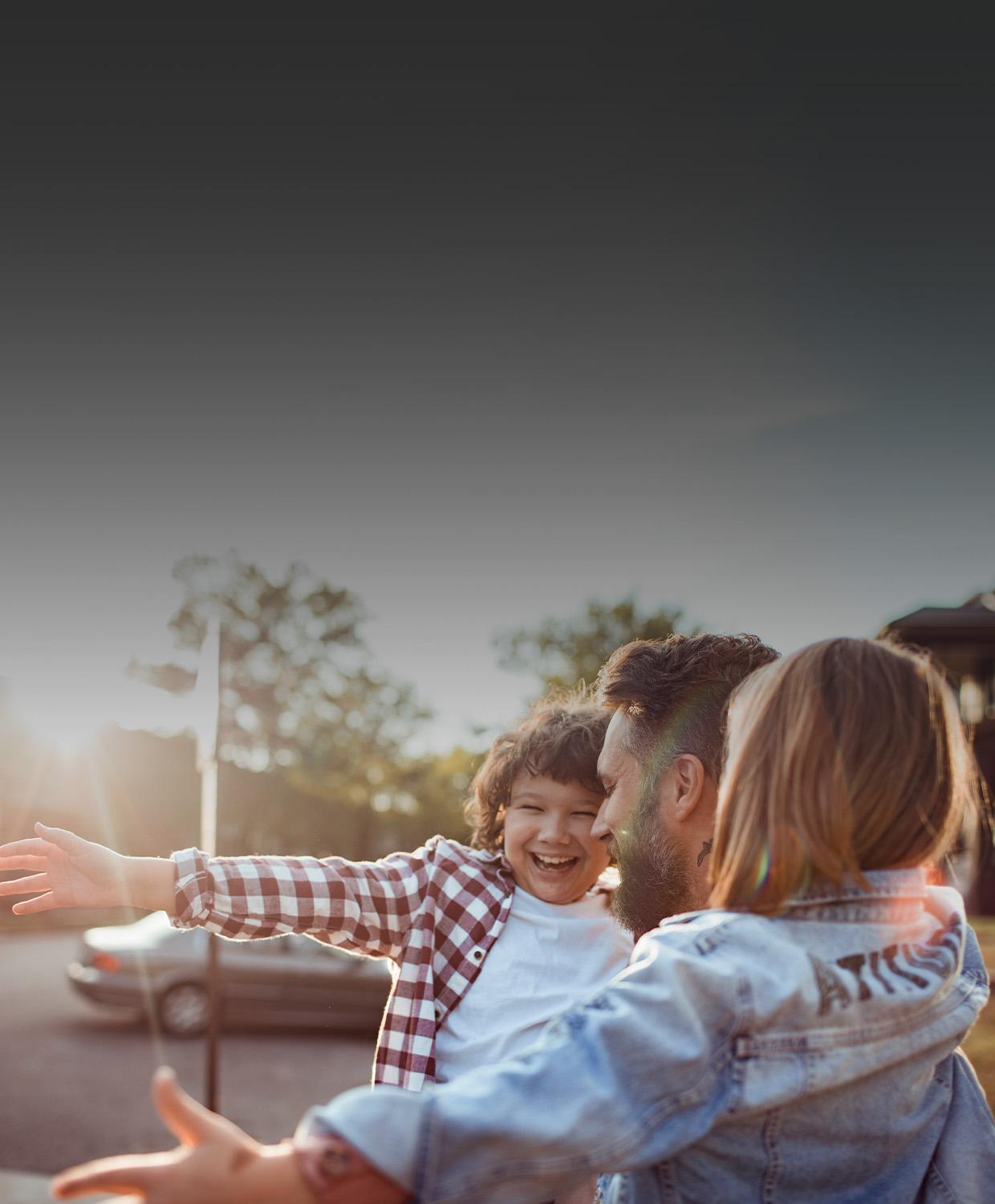 A man holds two children in his arms outdoors. They are smiling, and the children have their arms open wide.