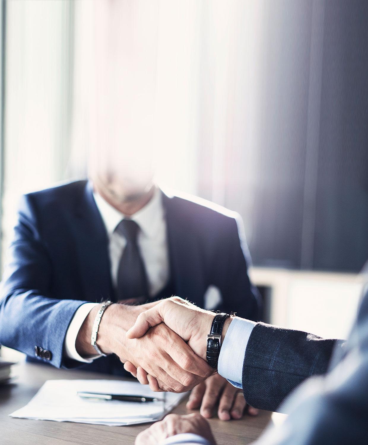 Two suited individuals shake hands across a conference room table.