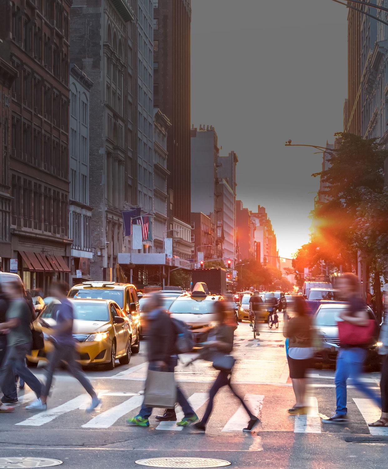 An image showing a vibrant community. A bright sunset in the background and people crossing a busy street at a crosswalk in the foreground.