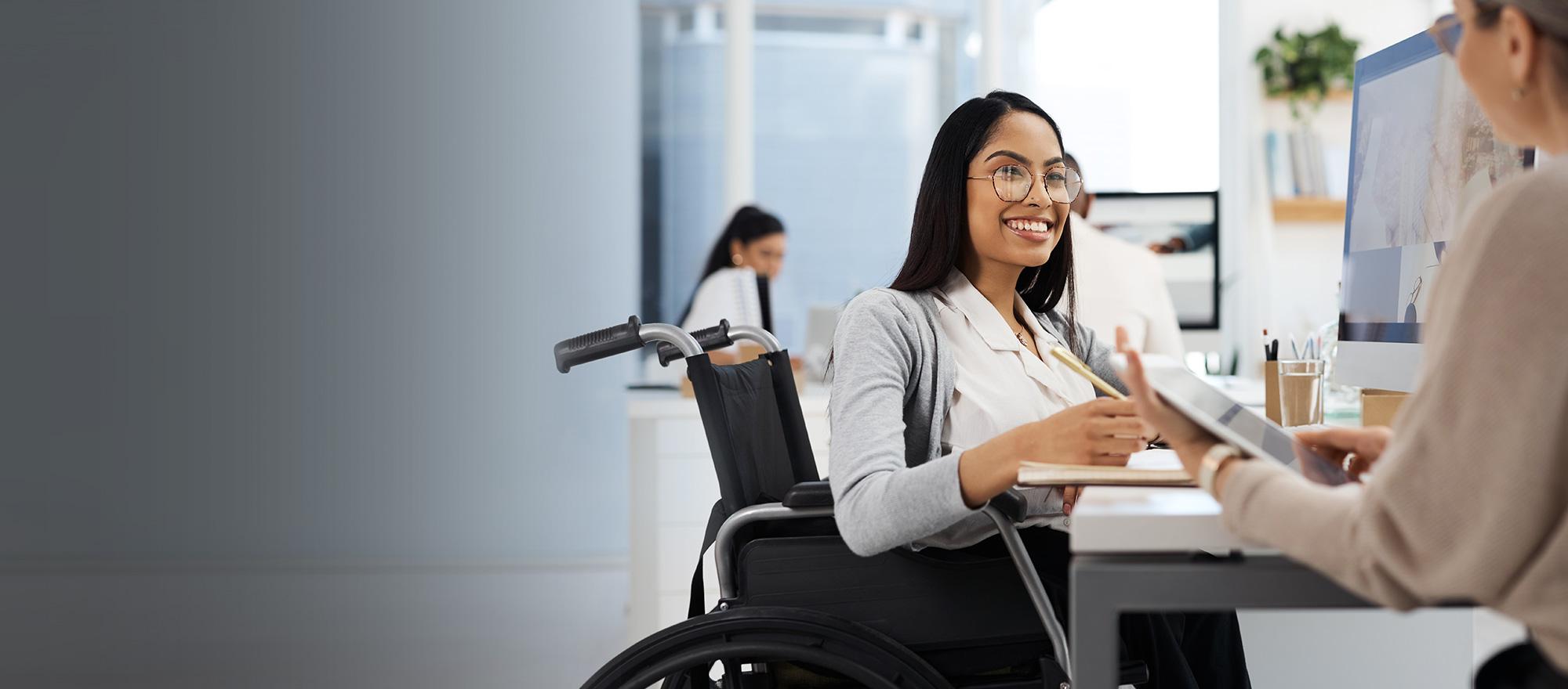 A woman in a wheelchair sits at a desk and smiles as she talks to another woman who holds a tablet in an office setting.