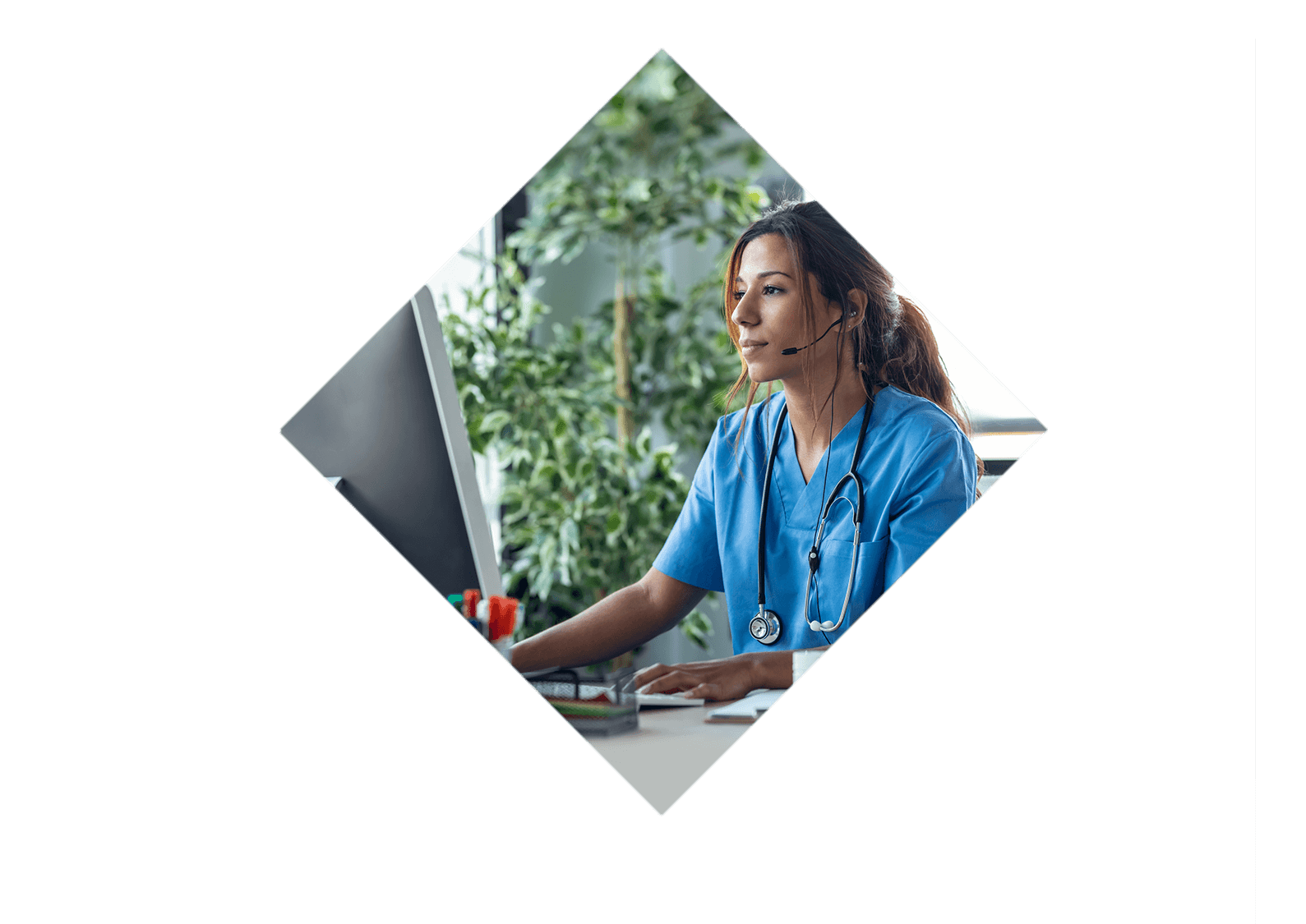 A woman in scrubs wearing a headset is typing on a desktop computer.