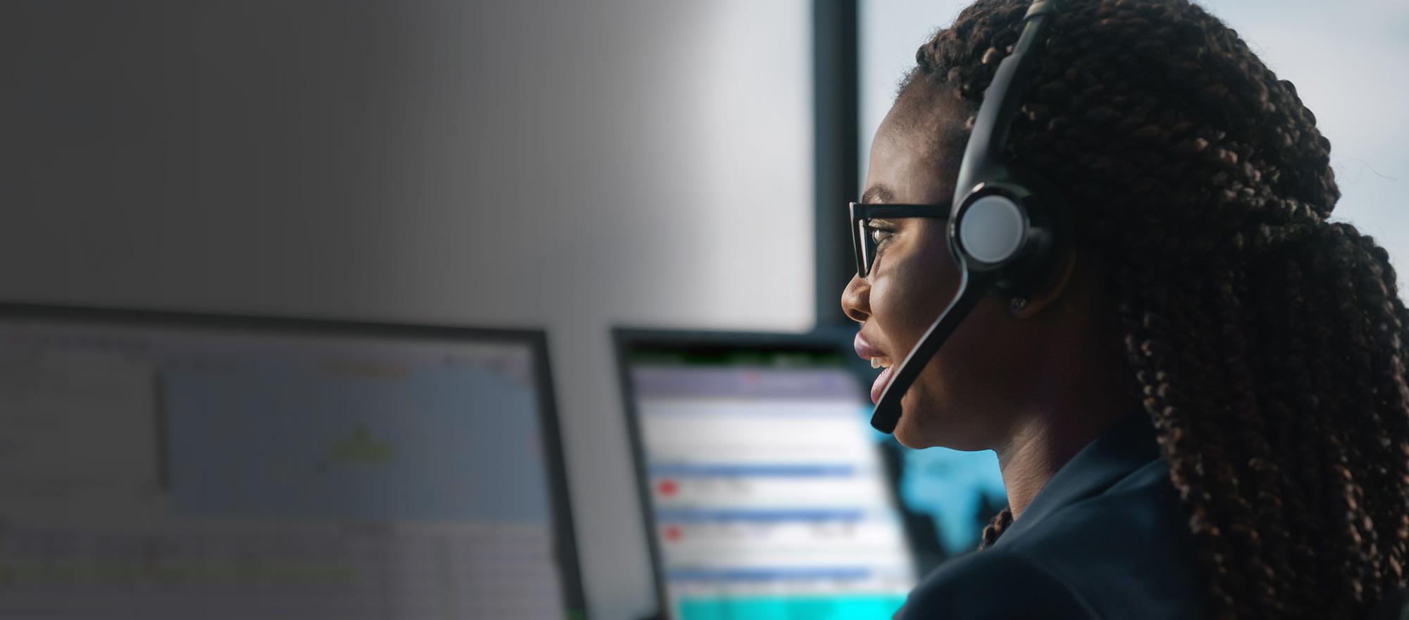 A mobile optimized photo of a female telecommunicator wearing a headset looking at a computer, smiling.