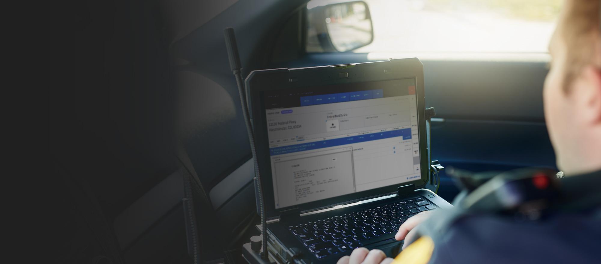 An over-the-shoulder view of a uniformed police officer interacting with software on a mobile laptop inside of a patrol car.