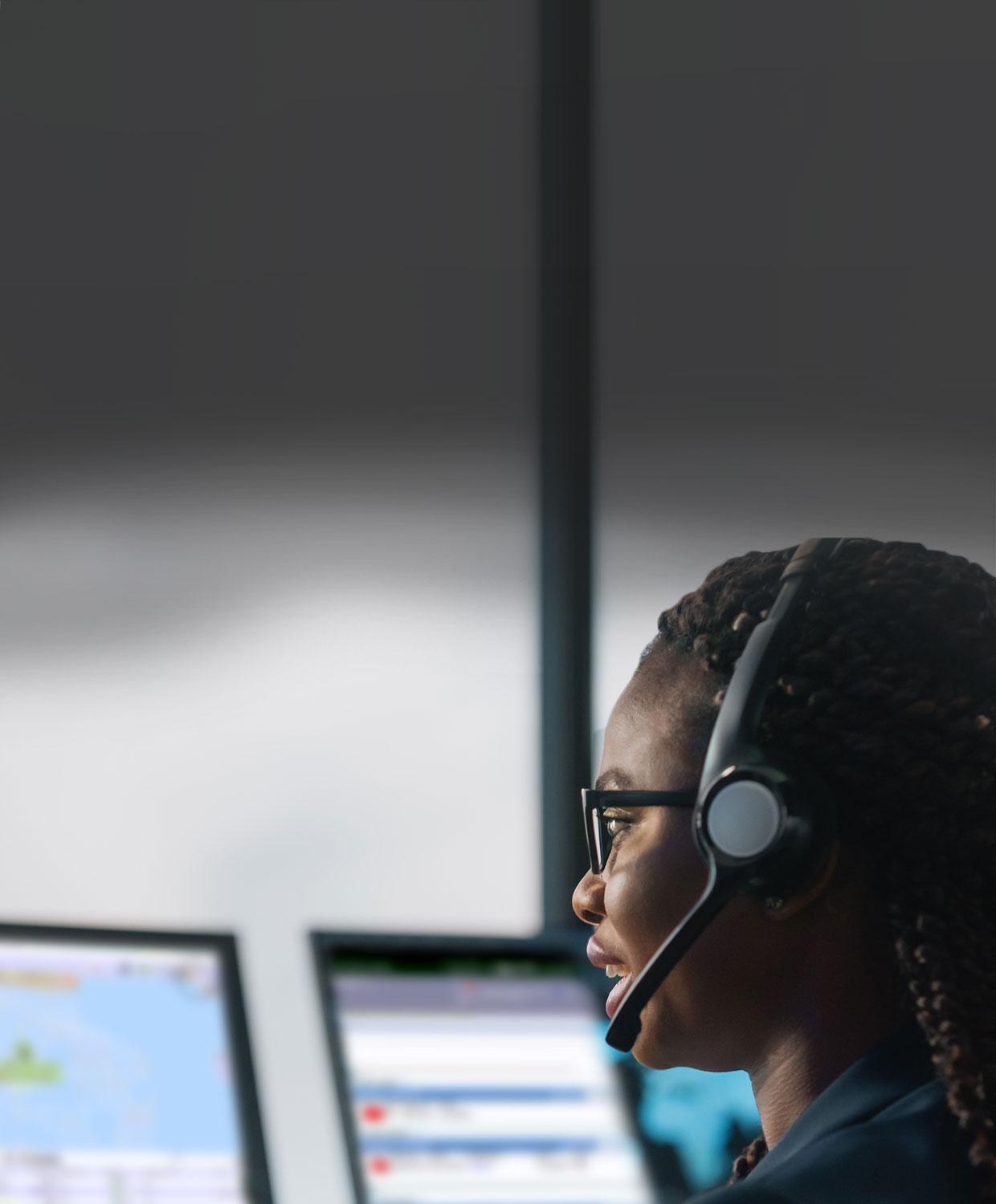 A mobile optimized photo of a female telecommunicator wearing a headset looking at a computer, smiling.