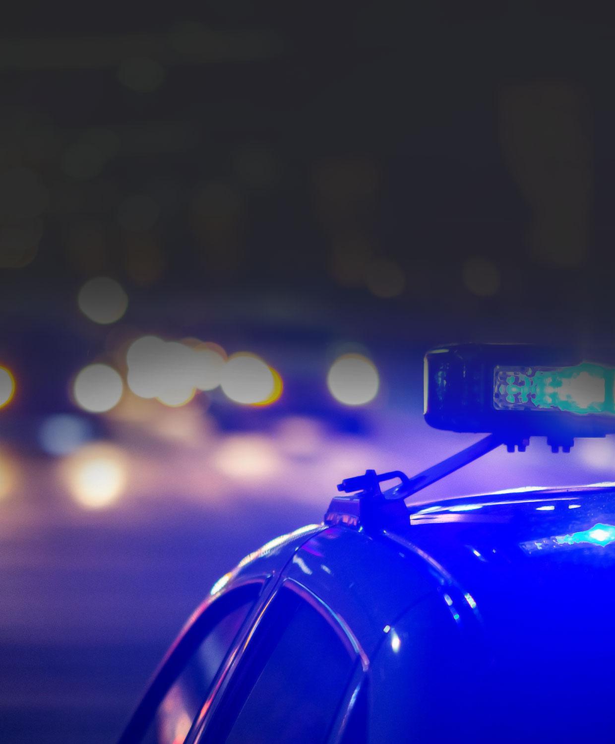 A dramatic photo of a patrol car on a city street at night with its emergency lights on. The car is in focus while the background is less distinguishable.