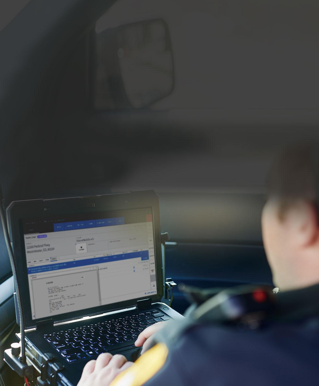 An over-the-shoulder view of a uniformed police officer interacting with software on a mobile laptop inside of a patrol car.