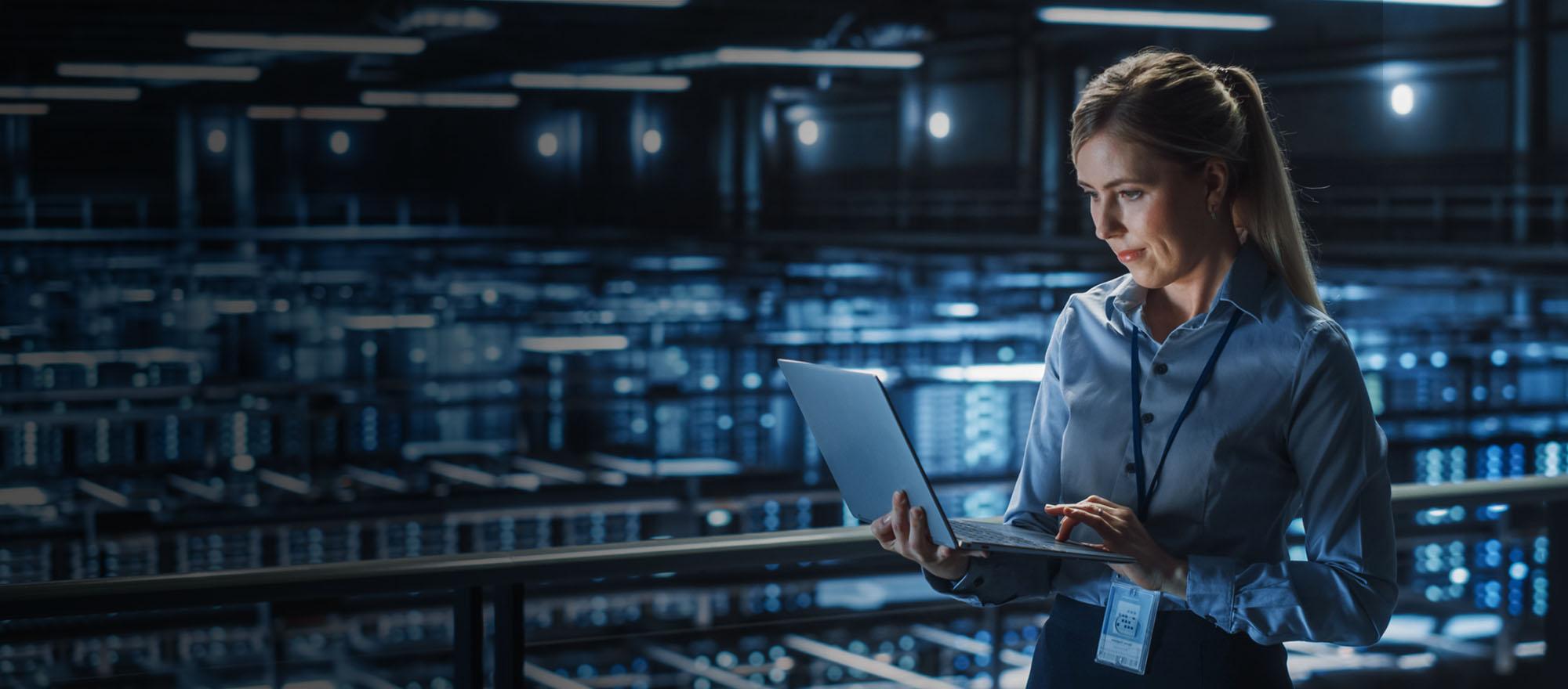 A woman in business causal clothing standing, holding a laptop, in an IT setting.